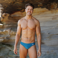 Man in blue green teal swimwear standing in front of a rock formation with water reflection, made in australia swimwear sydney